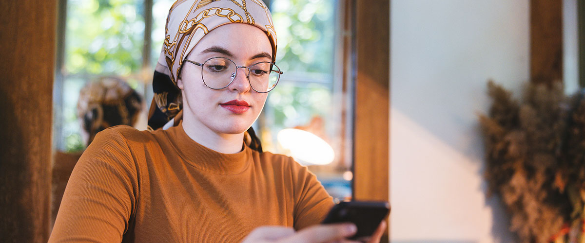 Young woman with glasses wearing a brown long sleeve shirt and headscarf, at home looking at her phone